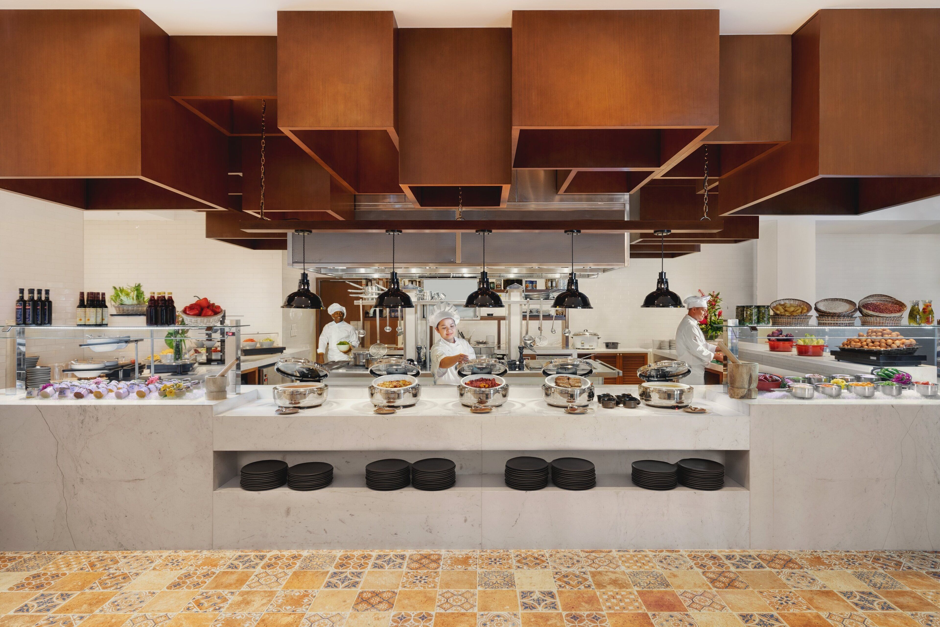 A modern kitchen with chefs preparing food, a buffet with various dishes, and a tiled floor.