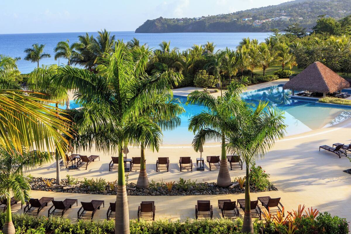 A resort with a pool, beach chairs, palm trees, and a view of the ocean.
