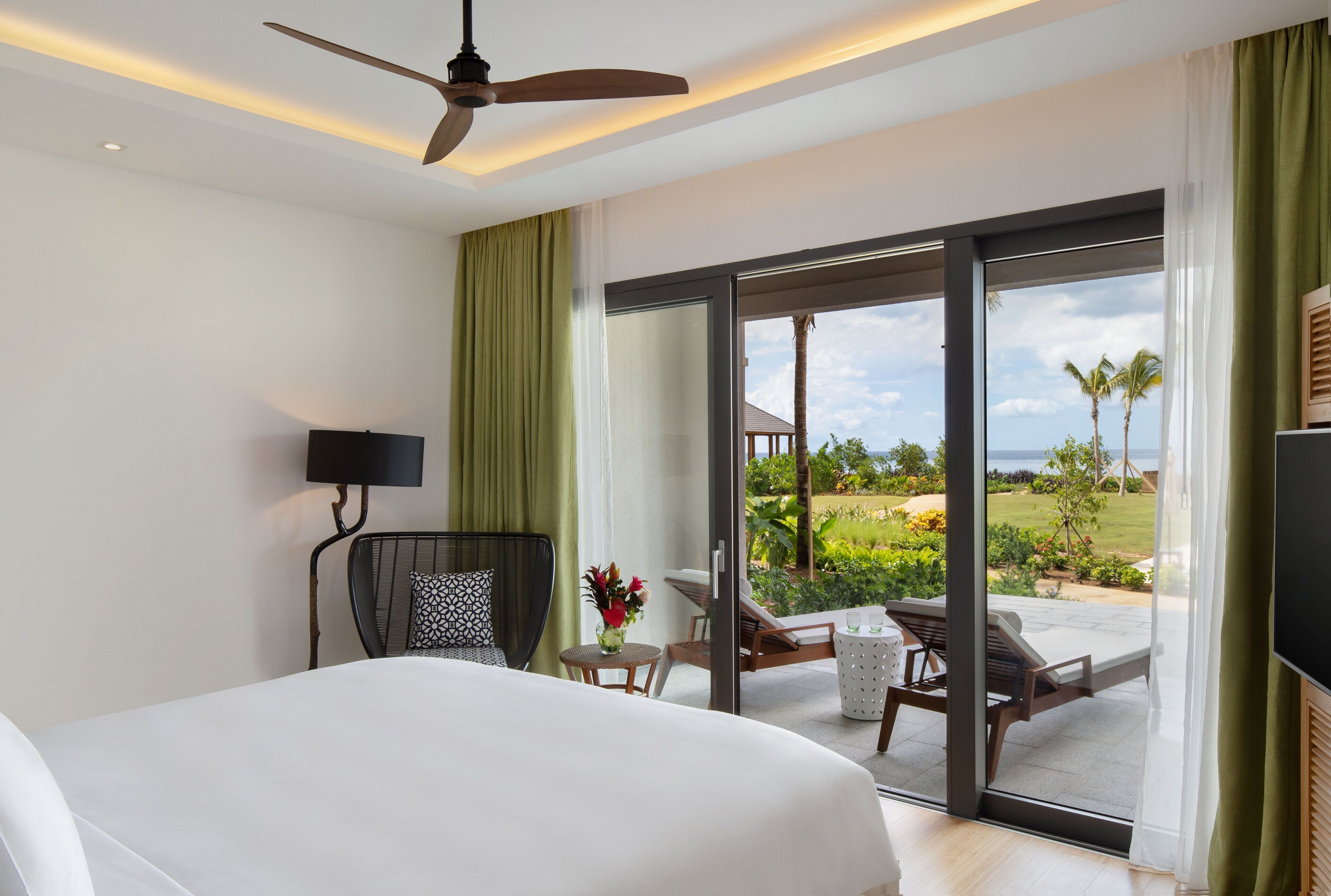 Habitación de hotel con cama, ventilador de techo, escritorio con lámpara, una silla, una mesita con un jarrón de flores y puerta corrediza de vidrio que da a un área exterior con piscina y vegetación.