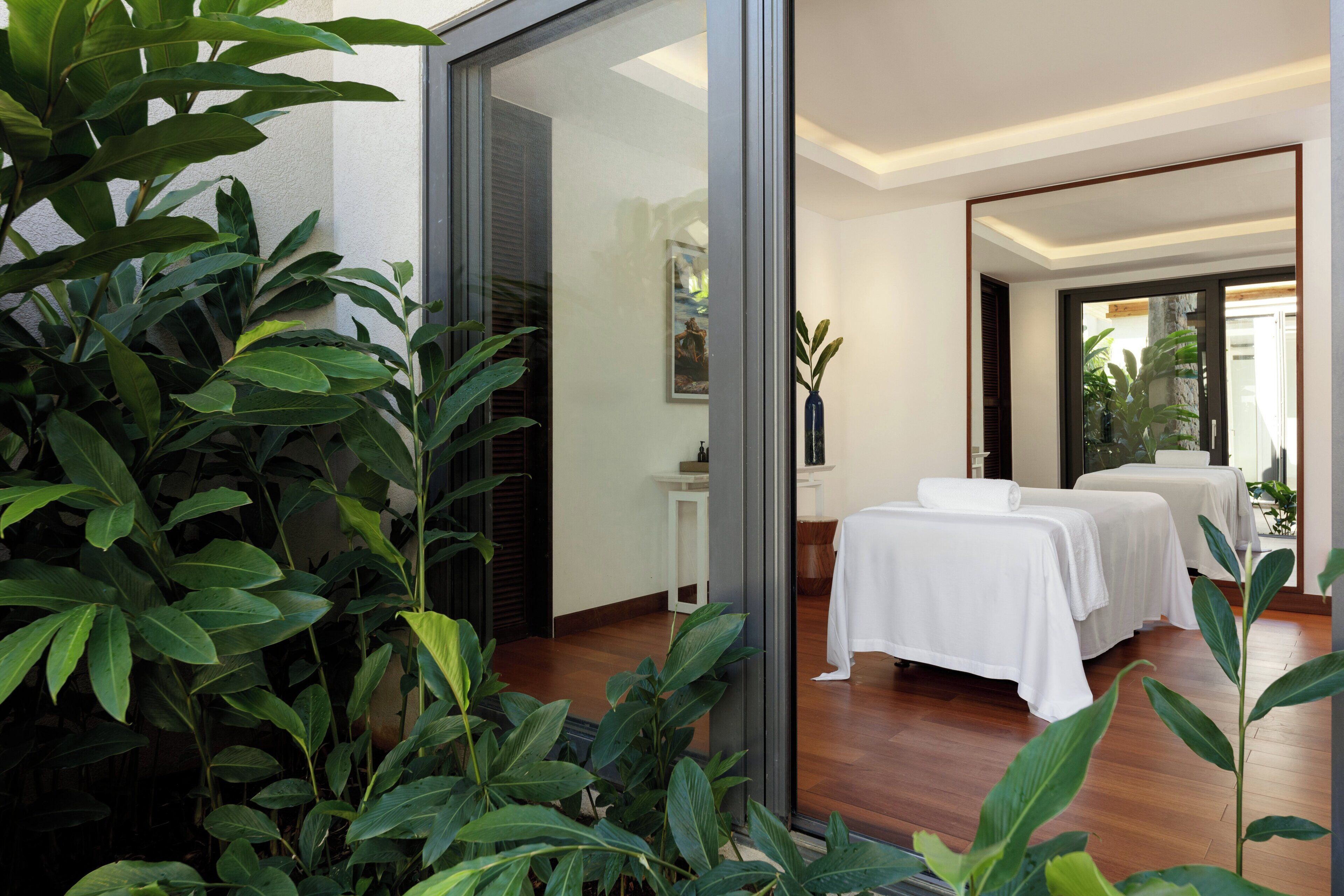 A spa room with two massage tables, a small white table with a vase, and a large window with a view of greenery.