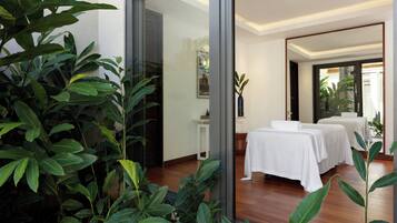 A spa room with two massage tables, a small white table with a vase, and a large window with a view of greenery.