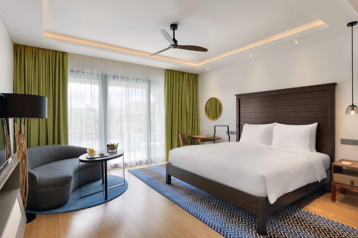 A modern hotel room with a large bed, a curved sofa, a small table, and a ceiling fan.