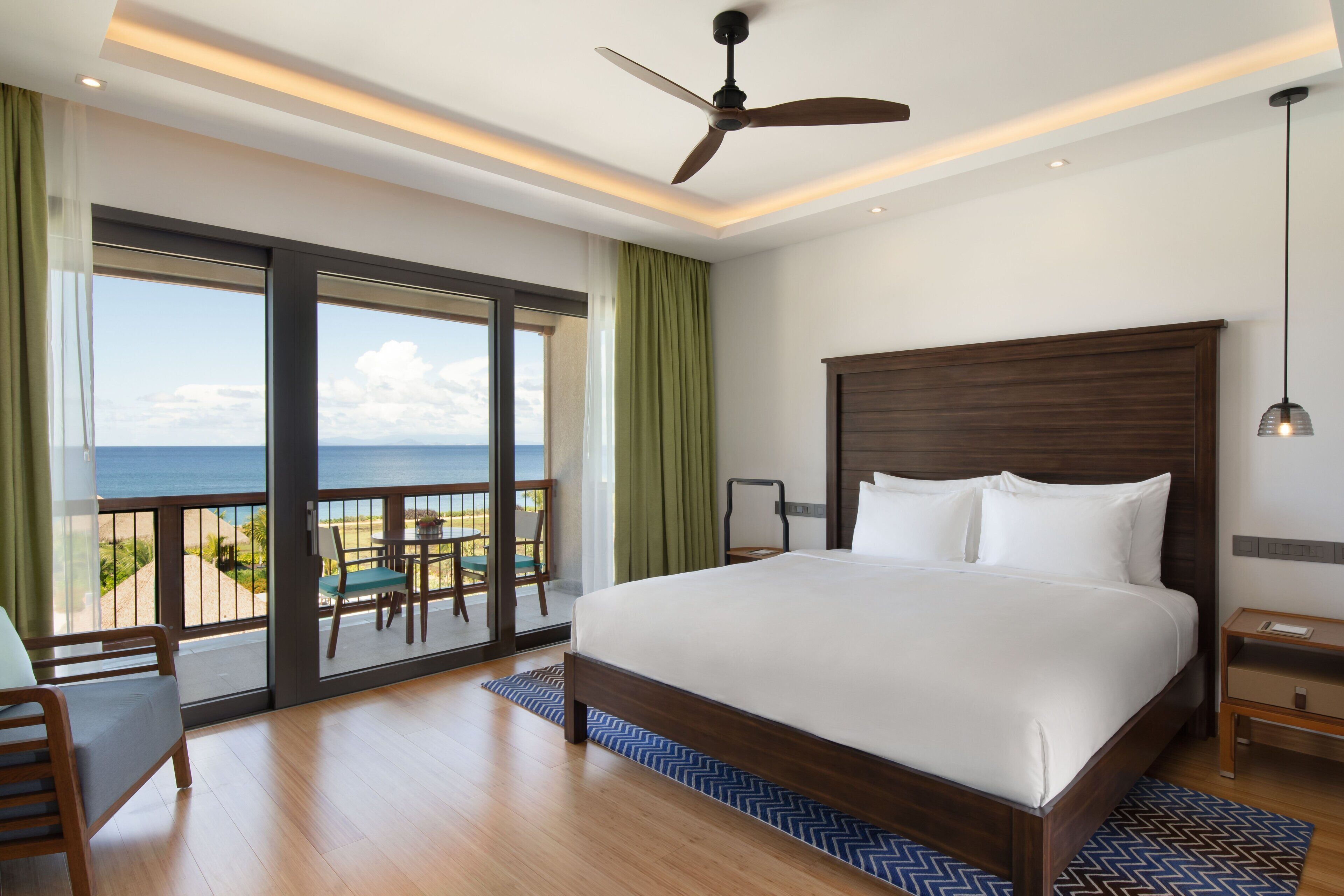 A spacious bedroom with a large bed, a ceiling fan, and a view of the ocean through a sliding glass door.
