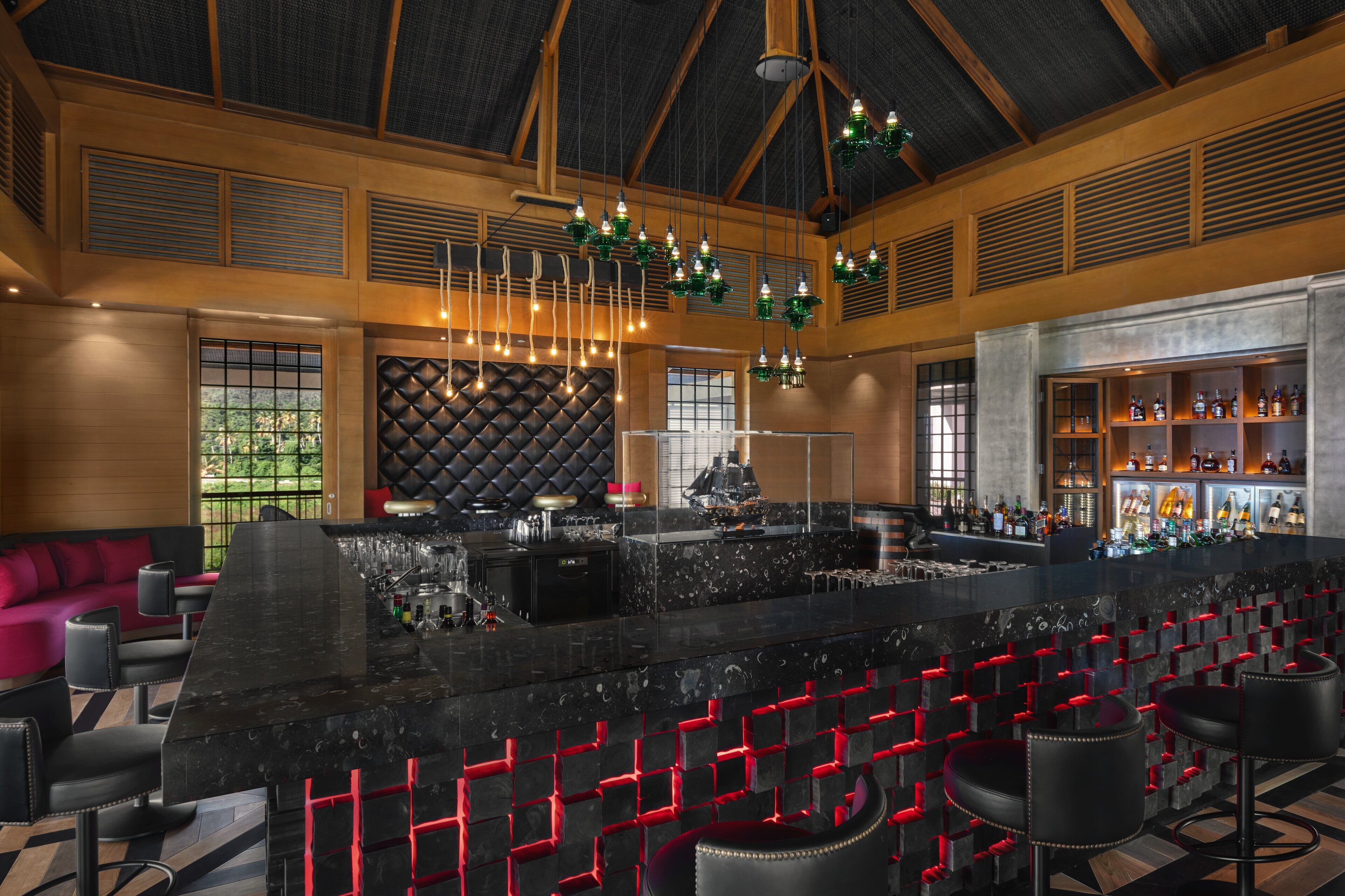 Uma área de bar com balcão de mármore preto, banquetas vermelhas e uma variedade de garrafas.