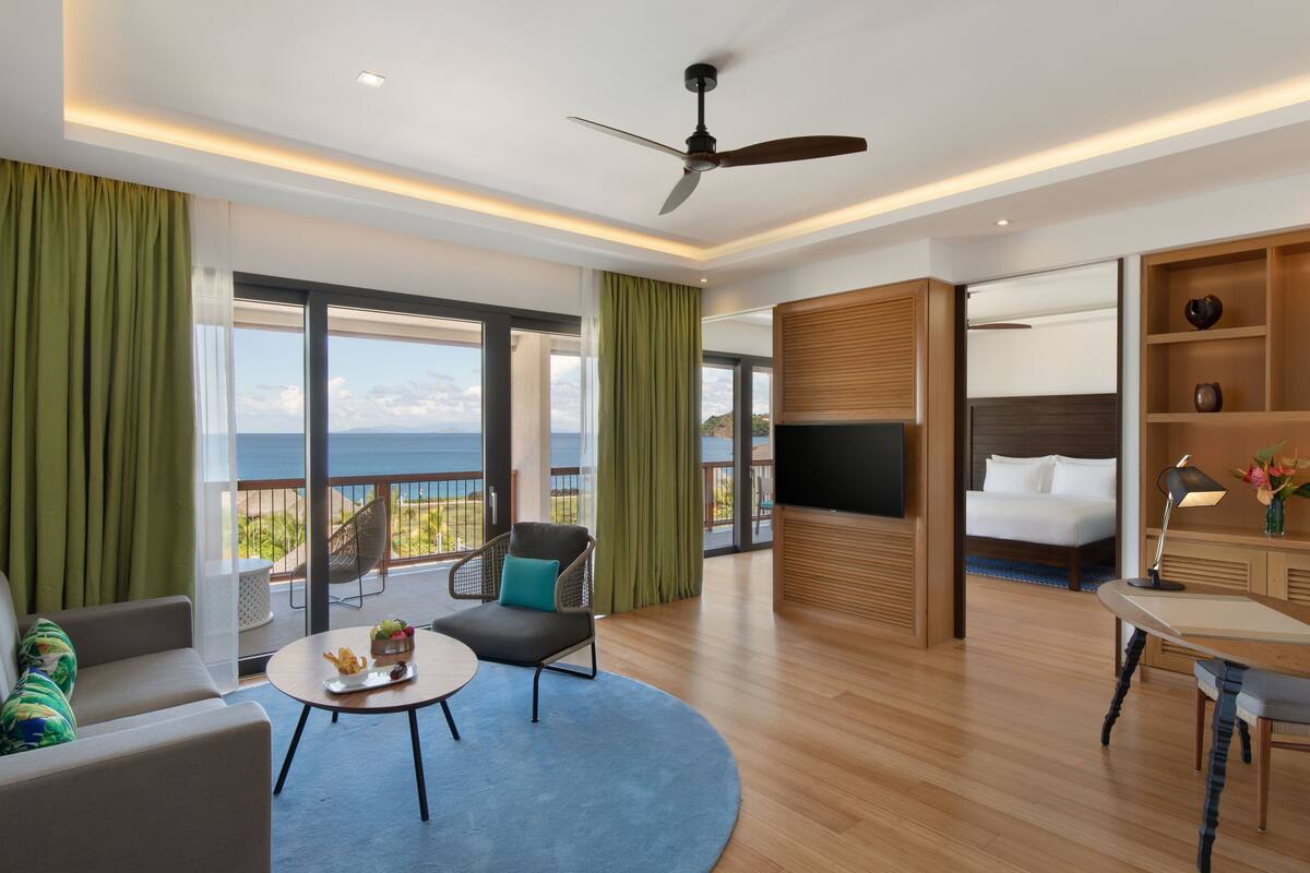 A modern hotel room with a balcony, a bed, a desk, and a seating area.