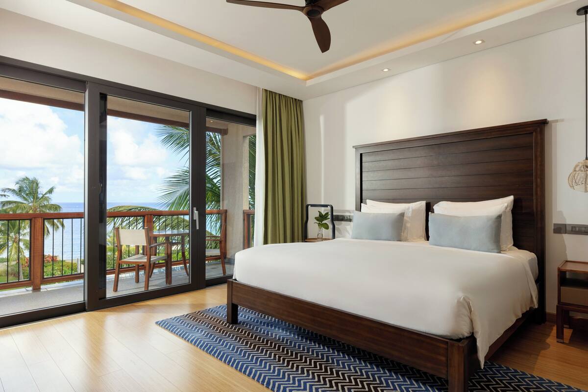 A spacious bedroom with a large bed, a ceiling fan, and a view of the ocean through a sliding glass door.
