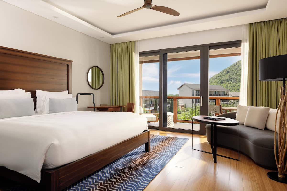 A modern hotel room with a large bed, a sofa, a small table, and a balcony with a view of a mountain.