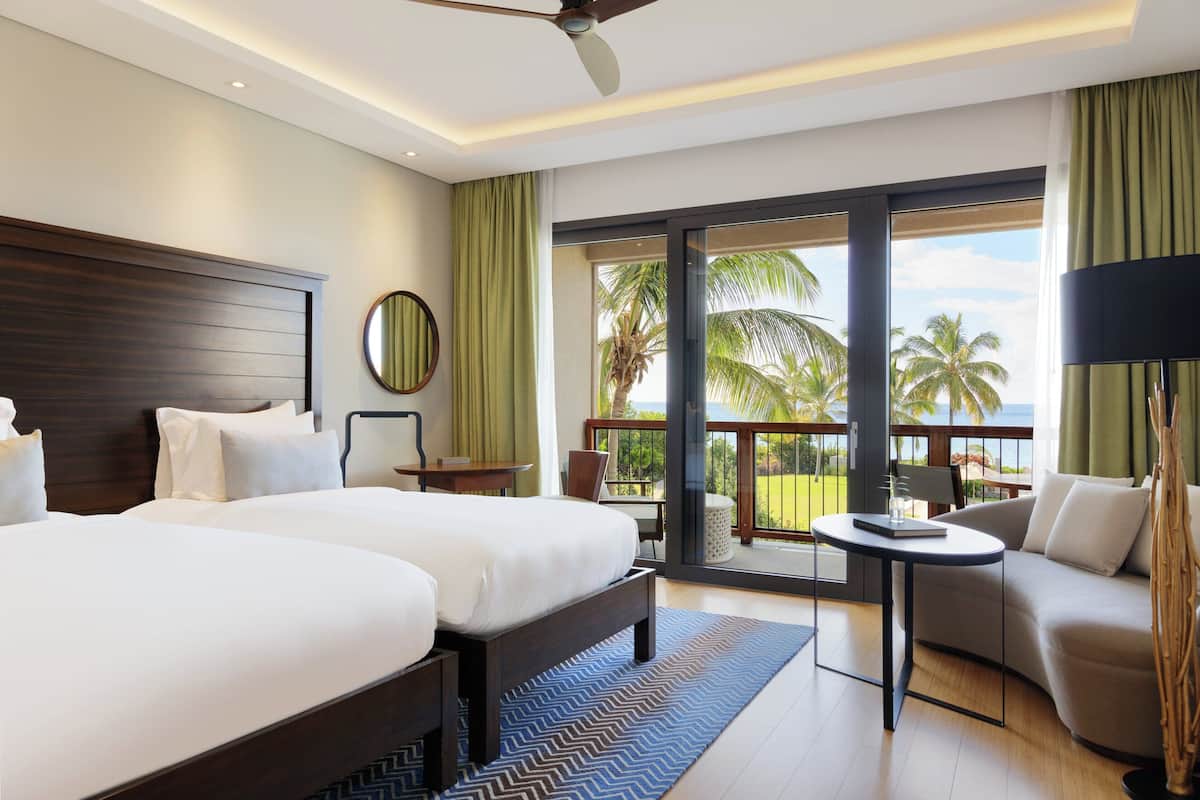 A hotel room with two beds, a balcony with a view of palm trees, and a seating area with a table and chairs.