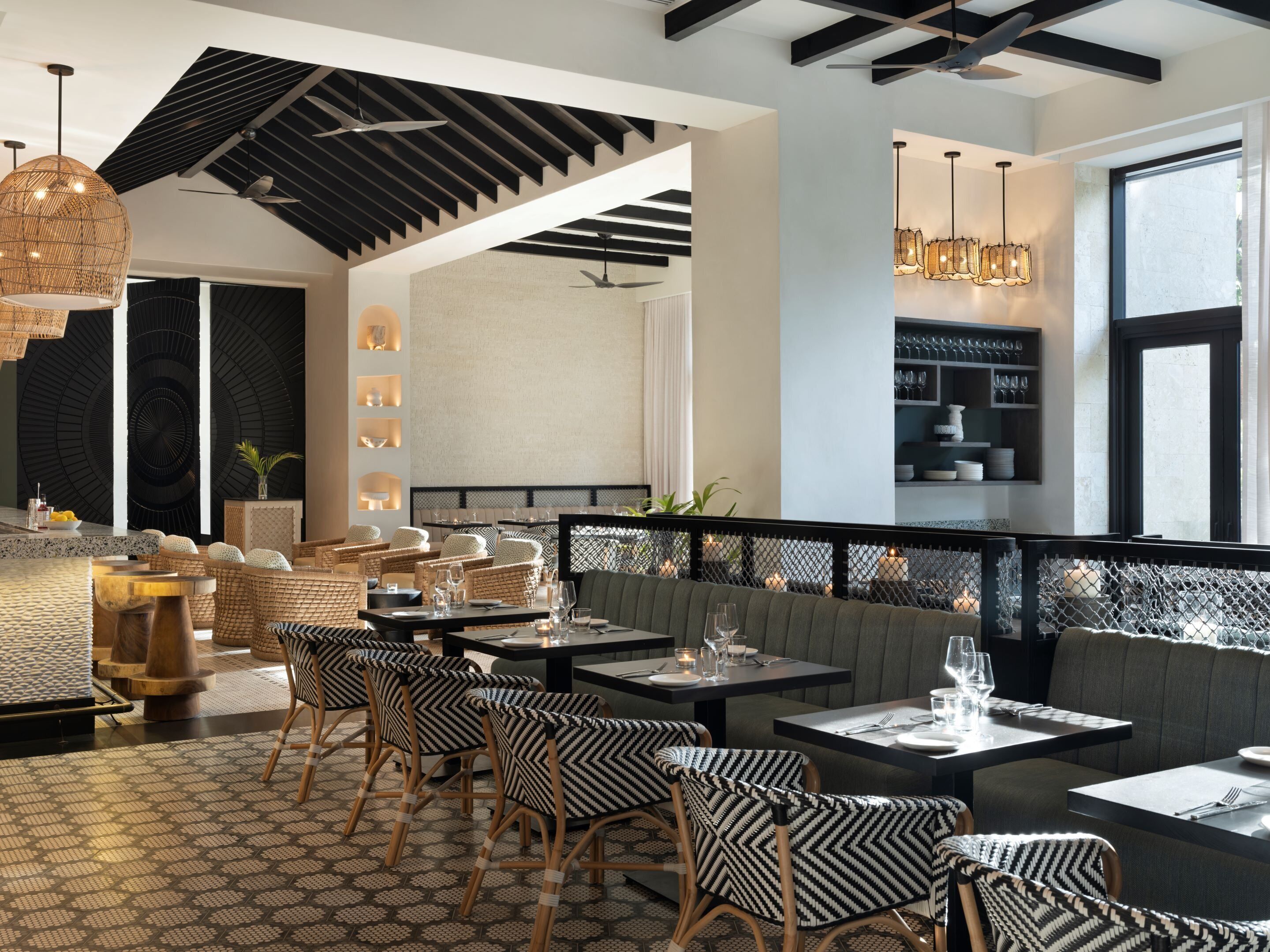 A restaurant interior with high ceilings, exposed beams, wicker furniture, and pendant lighting.