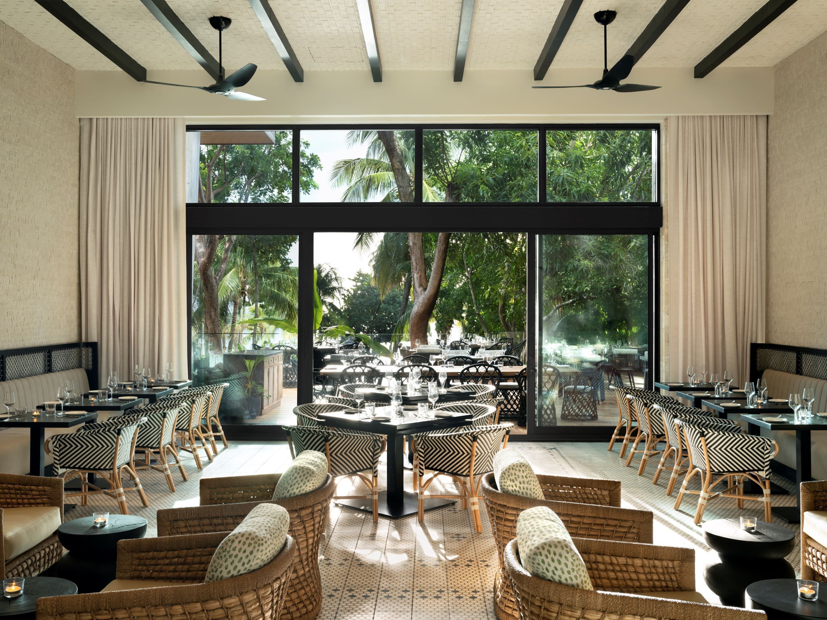 A restaurant with a large window view, multiple tables, wicker chairs, and ceiling fans.