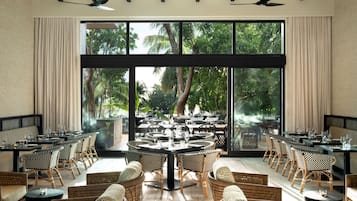 A restaurant with a large window view, multiple tables, wicker chairs, and ceiling fans.