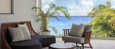 Un balcón con dos sillas de madera y una mesita pequeña, con vista al mar azul y palmeras.
