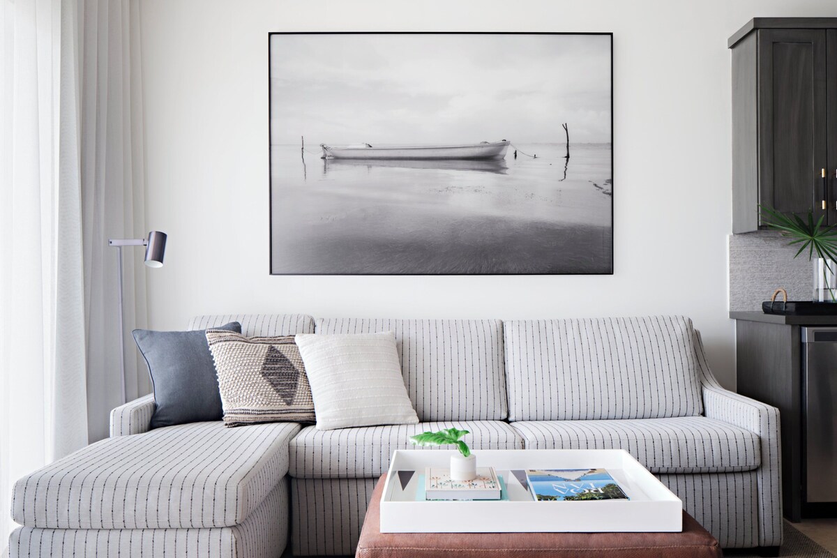 A modern living room with a striped sofa, a coffee table with books and a plant, and a large framed picture of a boat on water.