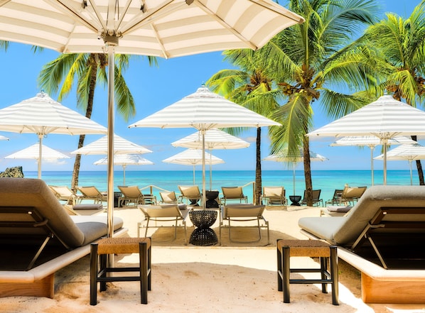 A beachside lounge area with multiple white-striped umbrellas, lounge chairs, and a view of the ocean.