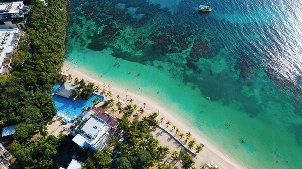 An aerial view of a beach resort with a swimming pool, palm trees, and clear turquoise waters. - Kimpton Grand Roatan Resort And Spa by IHG (Roatan)