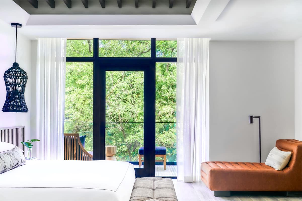 A modern bedroom with a large bed, a sofa, and a view of greenery through a large window.