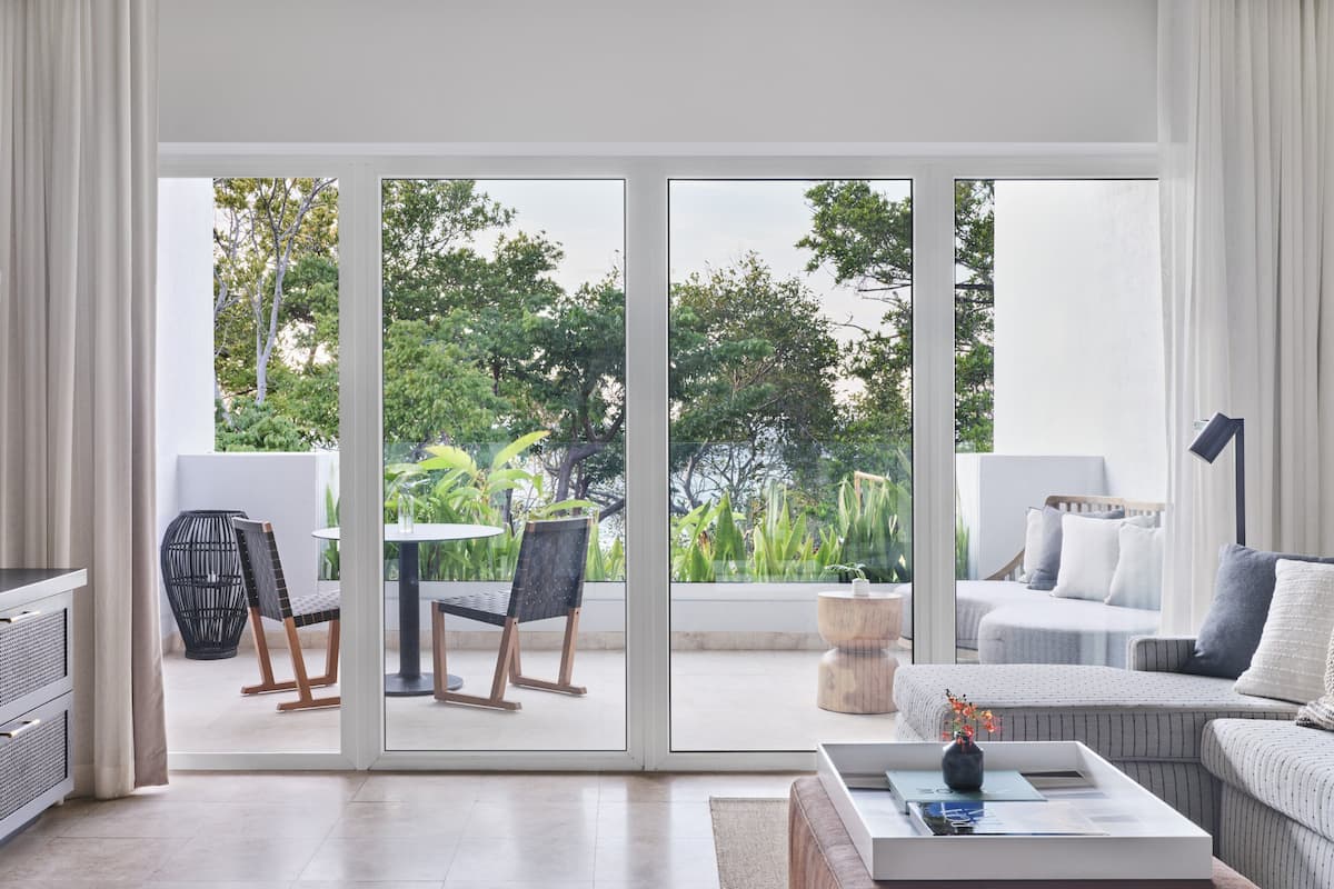 A modern living room with a sliding glass door leading to a garden. There is a sofa, a coffee table, and a dining area with chairs.