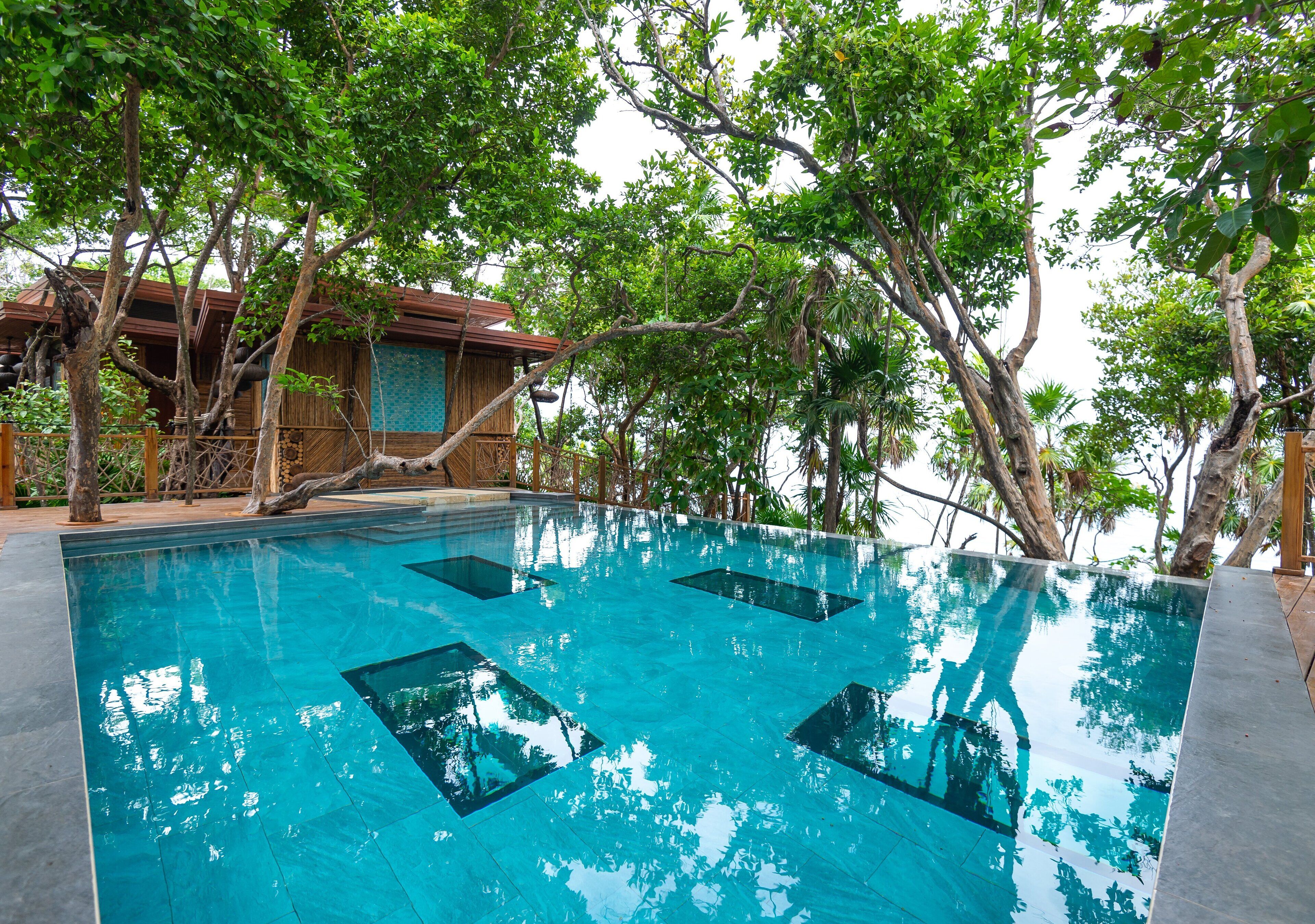 A clear swimming pool with a wooden deck surrounding it, trees in the background, and a building with a tiled wall.