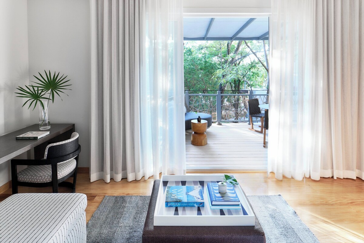 A modern living room with a sofa, a coffee table with books, a side table with a plant, and a view of an outdoor deck with a chair and table.