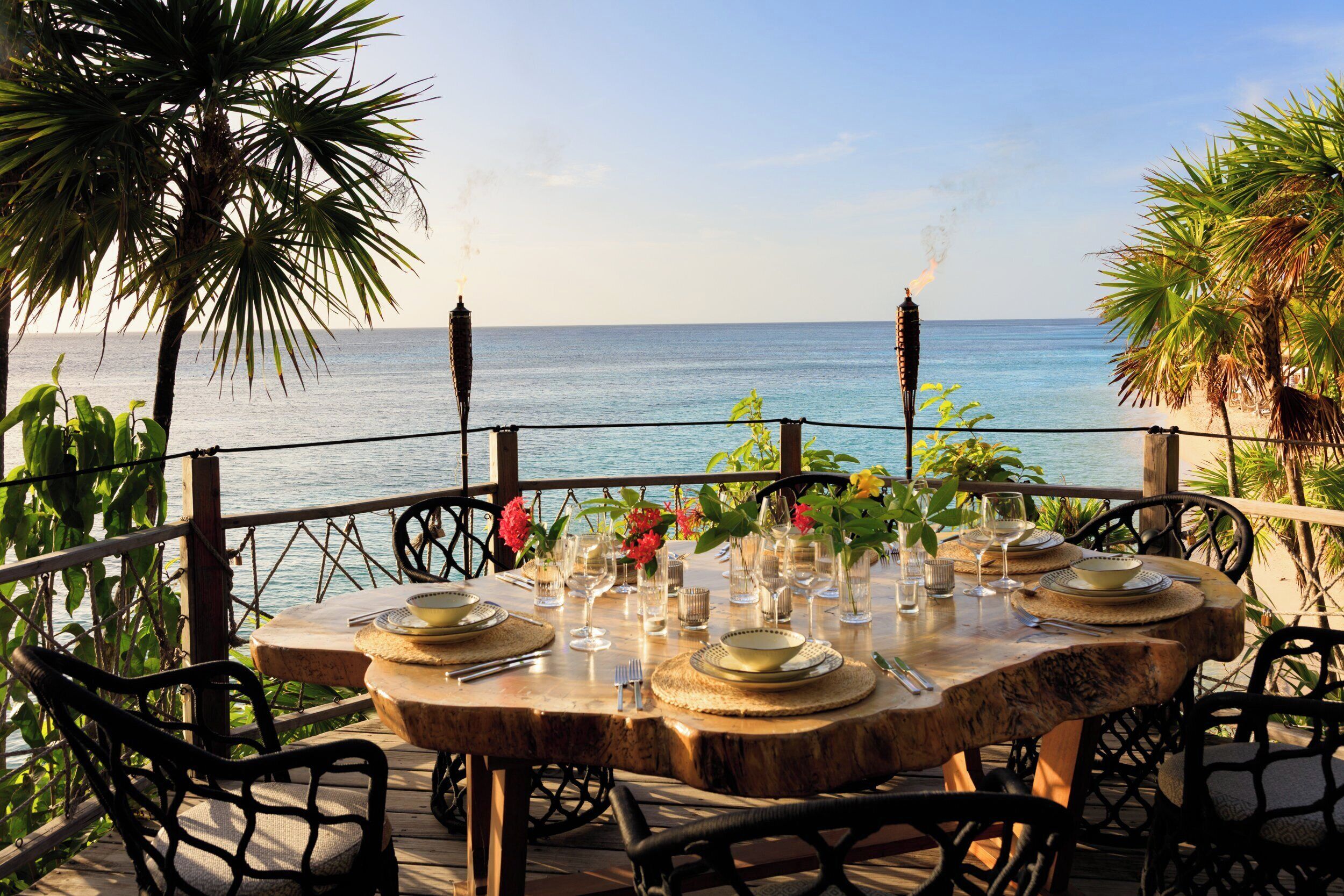 A table set for dining with a view of the sea, surrounded by chairs and palm trees.