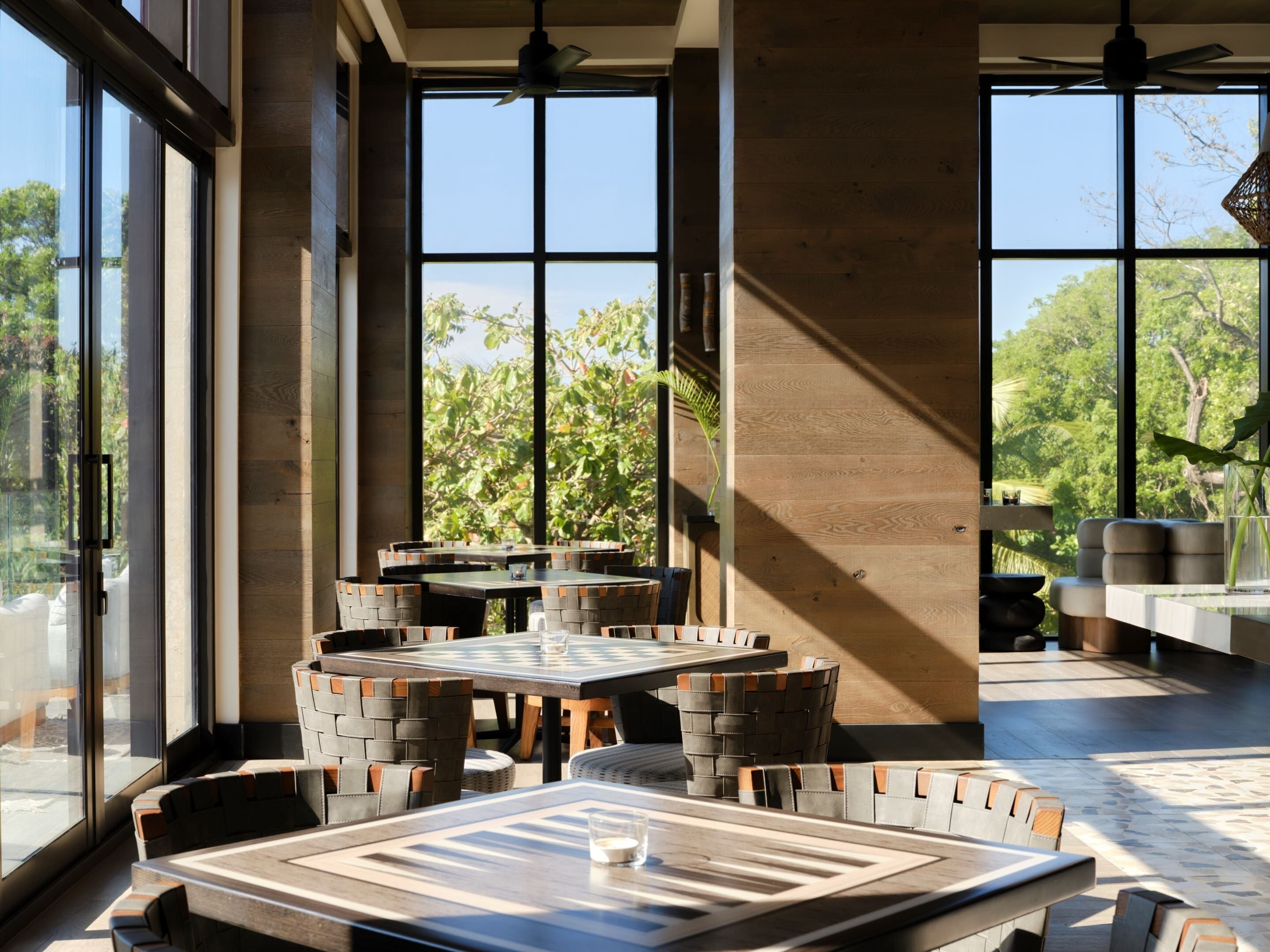 A modern outdoor dining area with large windows, wooden tables, and woven chairs.
