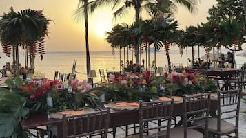 Un área de comedor al aire libre con mesas dispuestas para una comida, con vista al océano al atardecer.