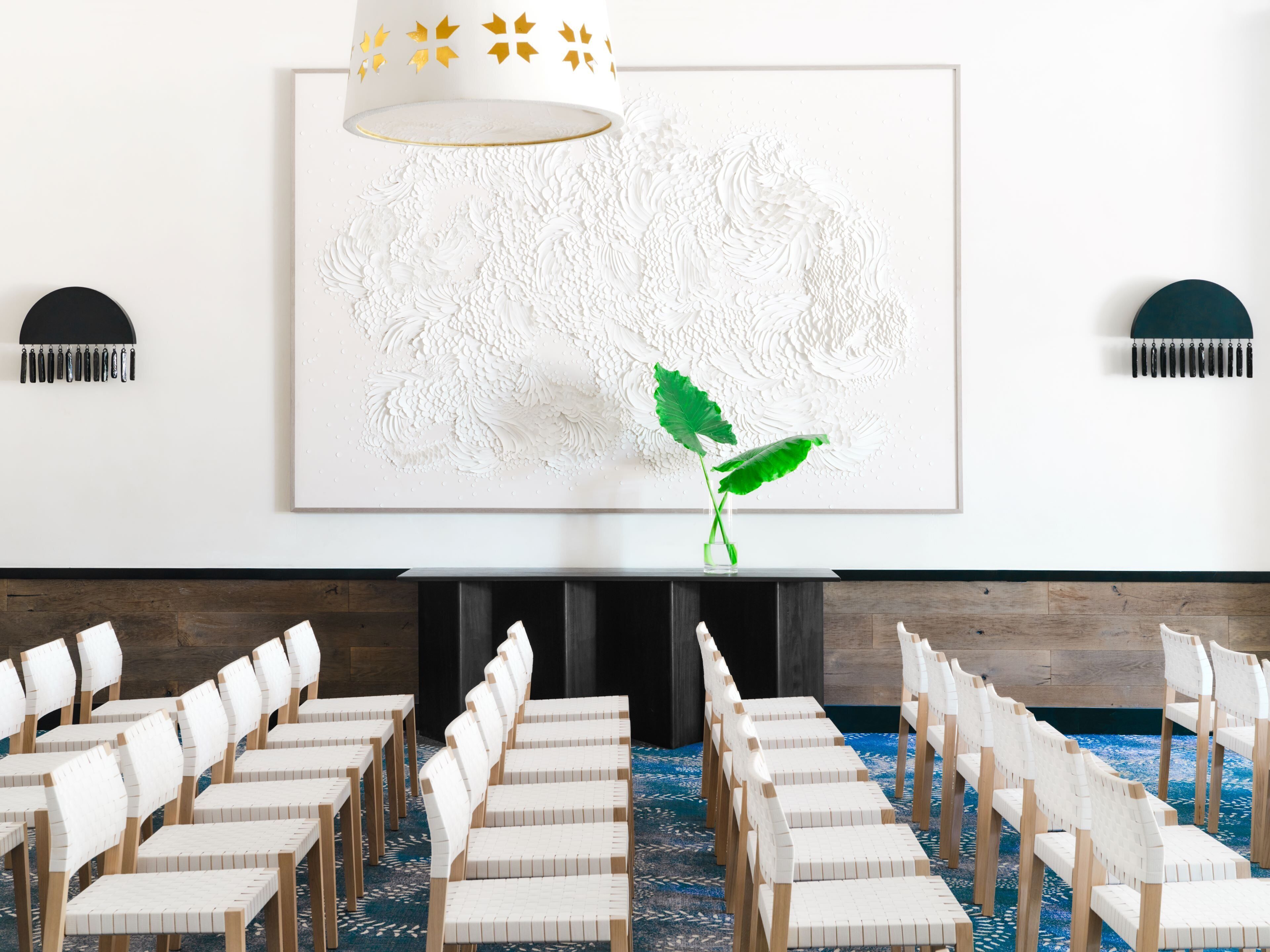Un salón de conferencias con sillas blancas, una obra de arte grande y texturizada en blanco, y una planta verde.