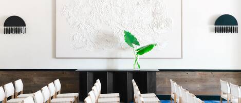Un salón de conferencias con sillas blancas, una obra de arte grande y texturizada en blanco, y una planta verde.