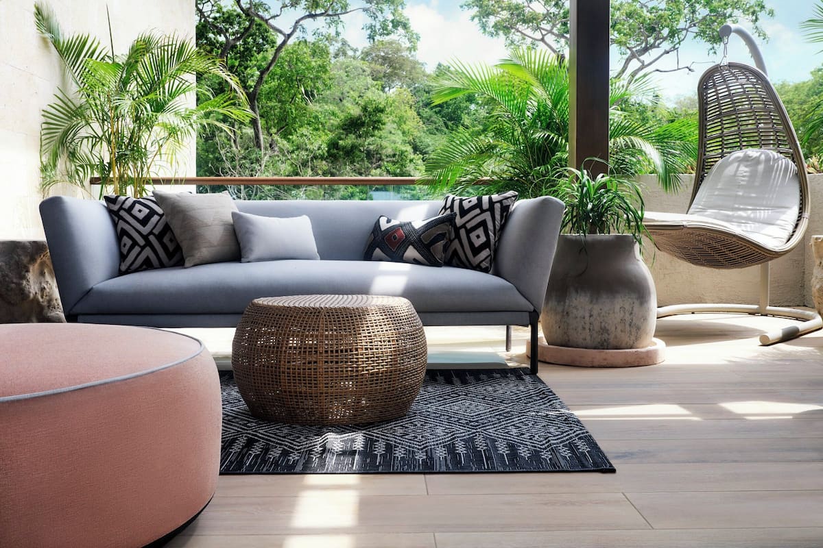 A modern outdoor seating area with a grey sofa, patterned cushions, a woven ottoman, and a wicker chair.