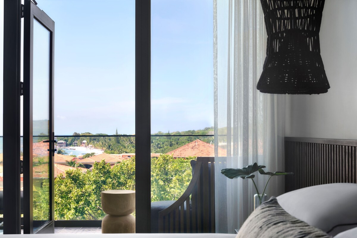 A bedroom with a large window, a bed, a chair, and a plant.