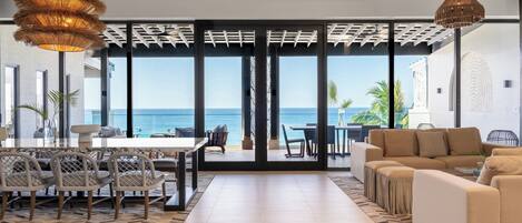 Een ruime woonkamer met een groot raam met uitzicht op zee, een eethoek met rieten stoelen, en een beige bank.