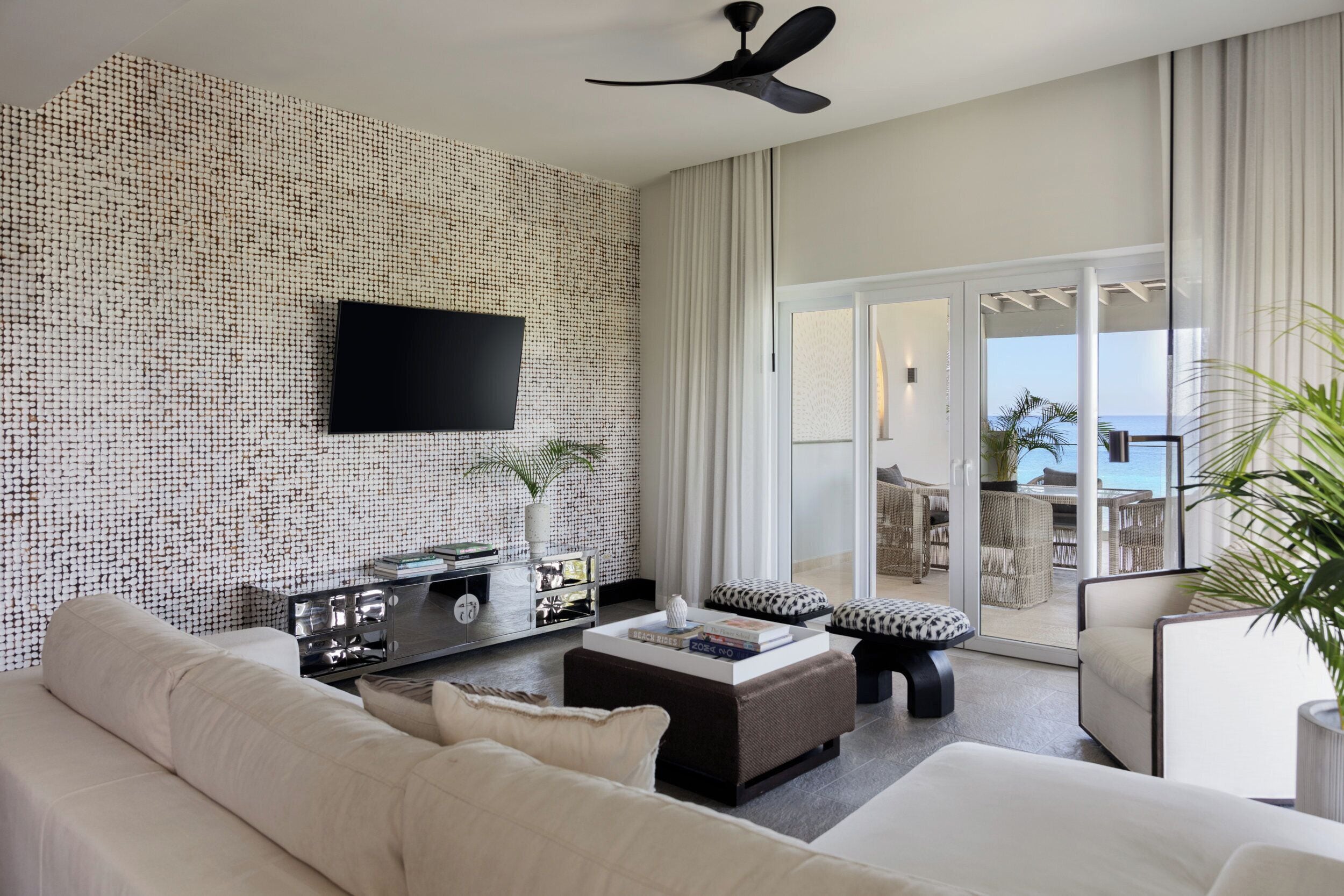 A modern living room with a flat-screen TV mounted on a textured wall, a ceiling fan, a large window with curtains, a sofa, a coffee table, and a view of the sea through the glass door.