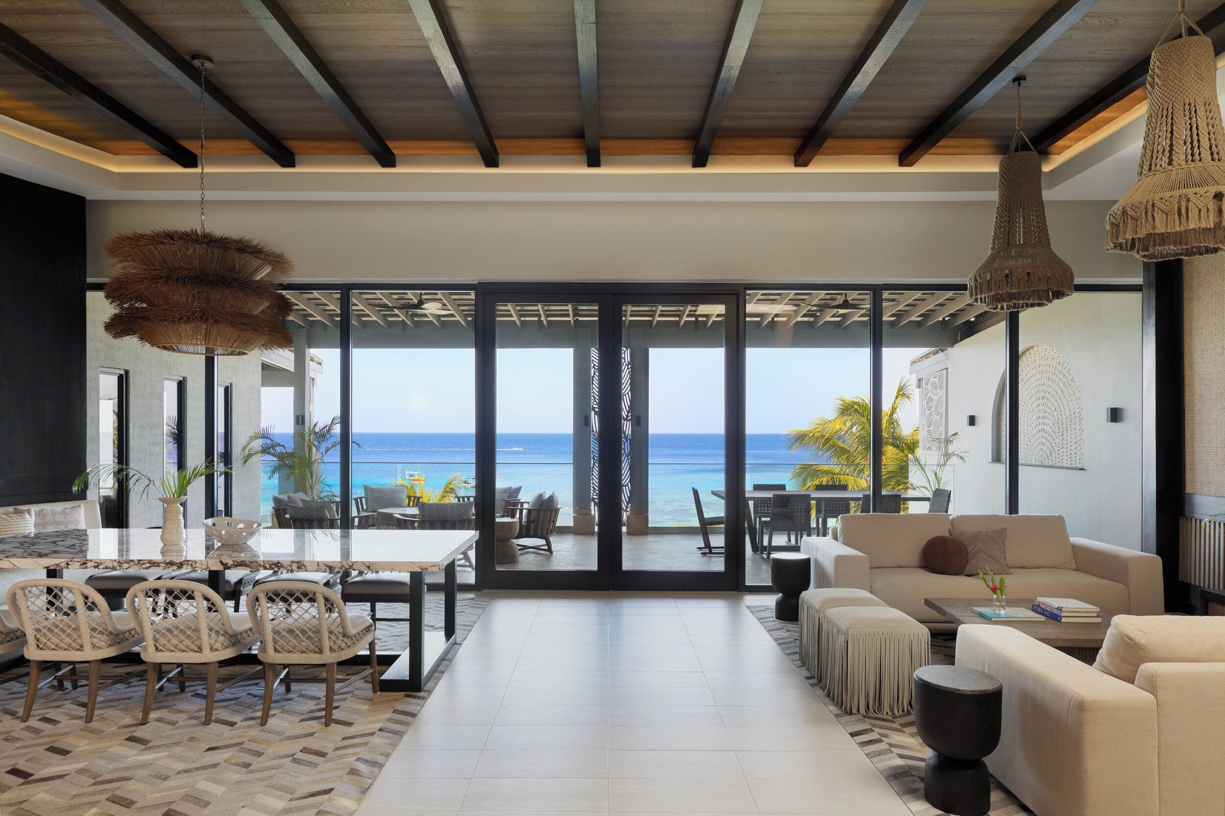 A spacious living area with a large glass door leading to a balcony overlooking the ocean. There are multiple seating areas, including a sofa set and a dining table with chairs.