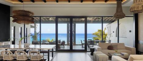 A spacious living area with a large glass door leading to a balcony overlooking the ocean. There are multiple seating areas, including a sofa set and a dining table with chairs.