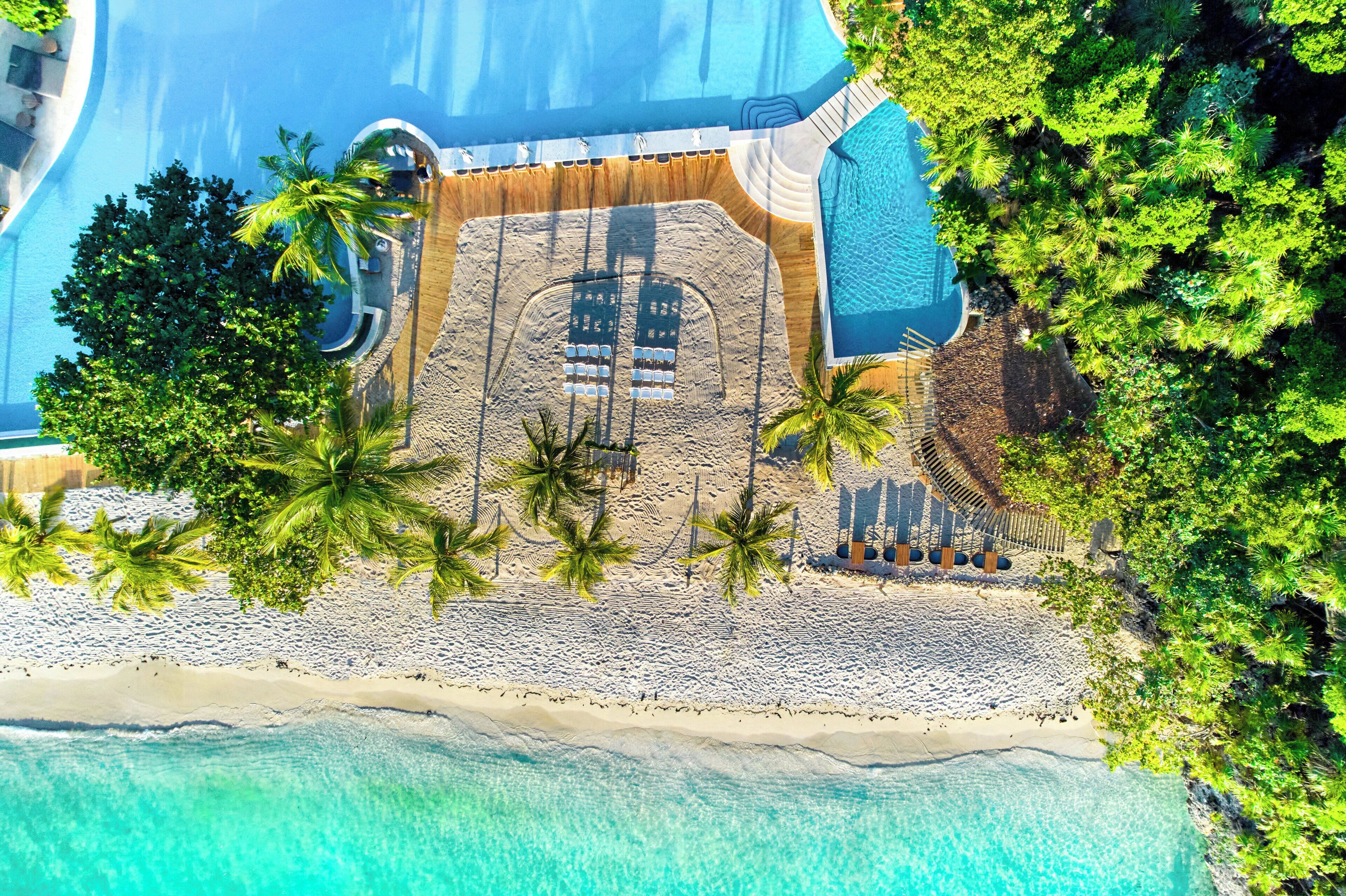 An aerial view of a resort with a large swimming pool, a sandy beach area, and palm trees.