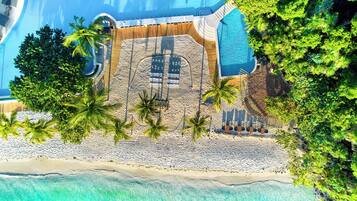 An aerial view of a resort with a large swimming pool, a sandy beach area, and palm trees.