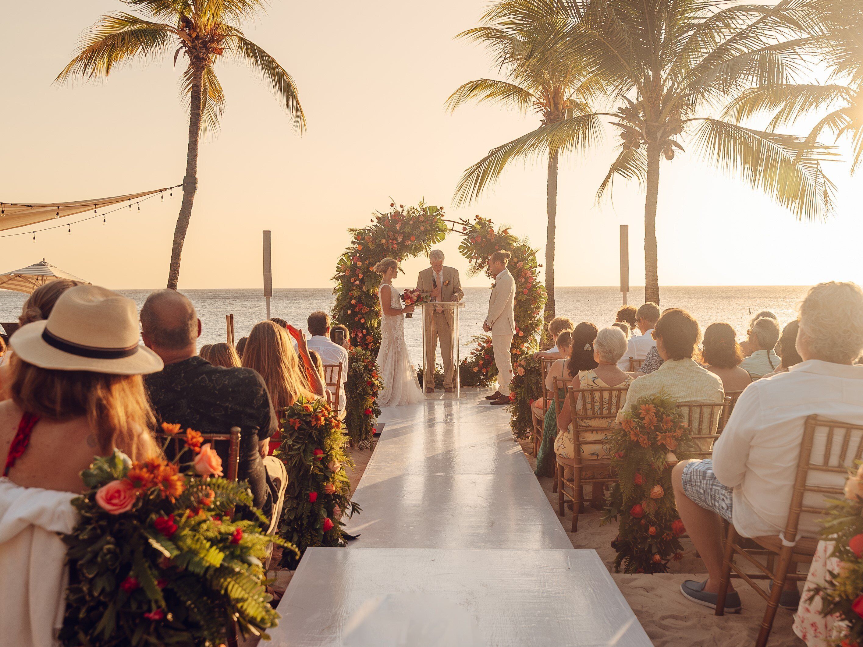 A beachside wedding ceremony with a floral arch and guests seated on chairs.