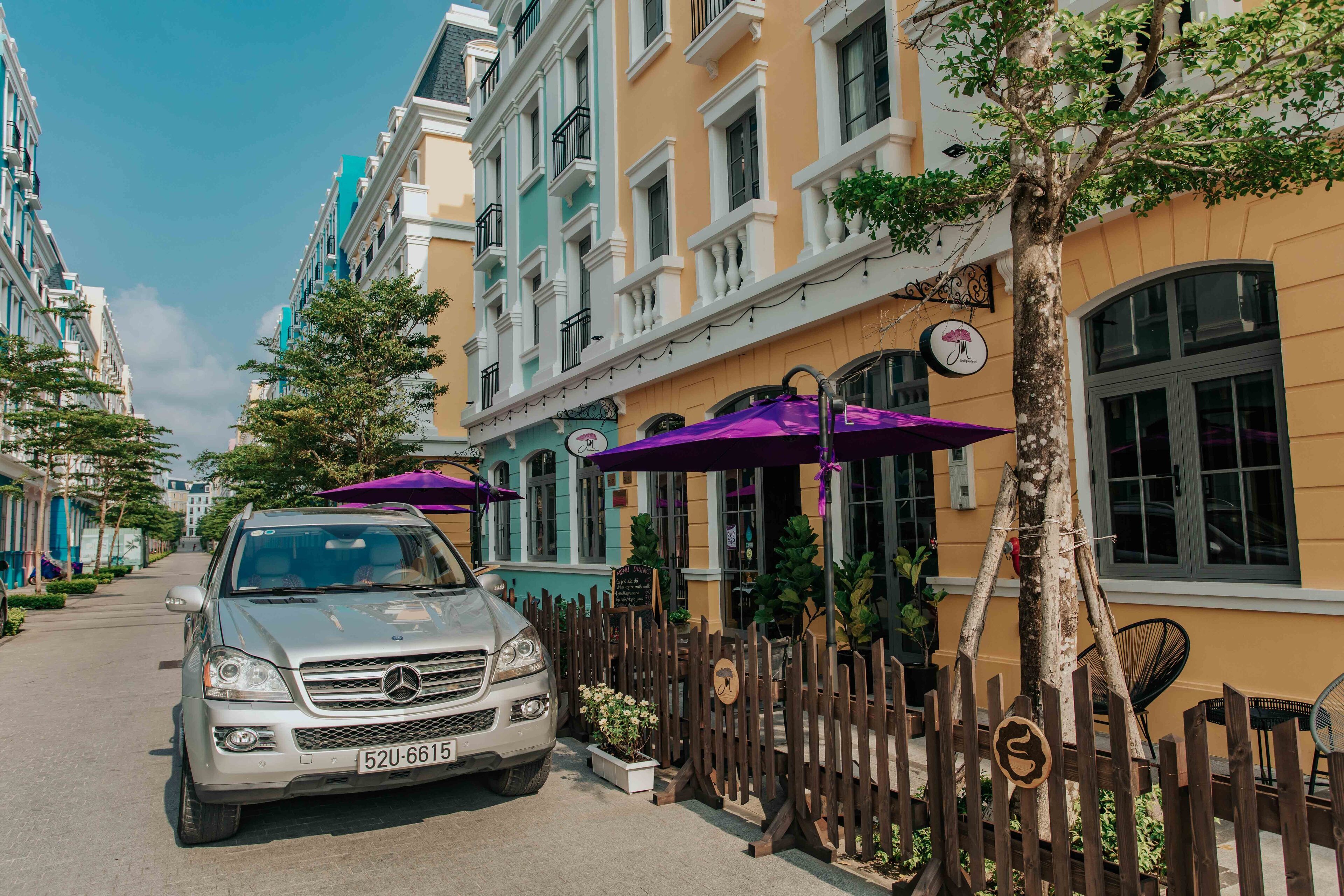 Une voiture argentée est garée dans une rue bordée d’immeubles aux couleurs vives, d’une clôture en bois et de parasols d’extérieur.
