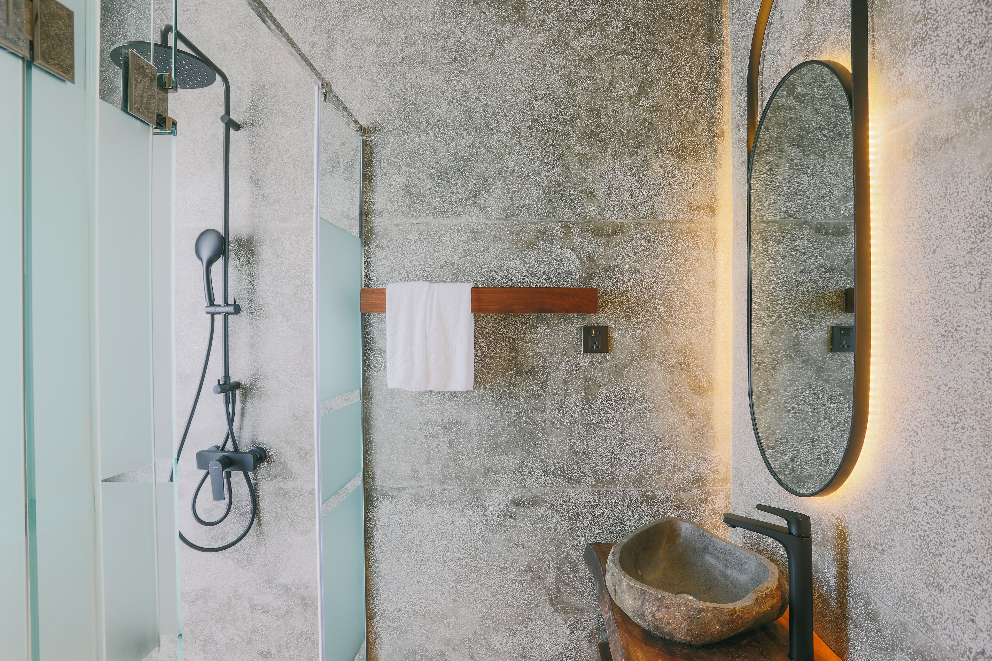 A bathroom with a concrete wall, a wooden shelf with a white towel, a stone basin, and a mirror with lighting.