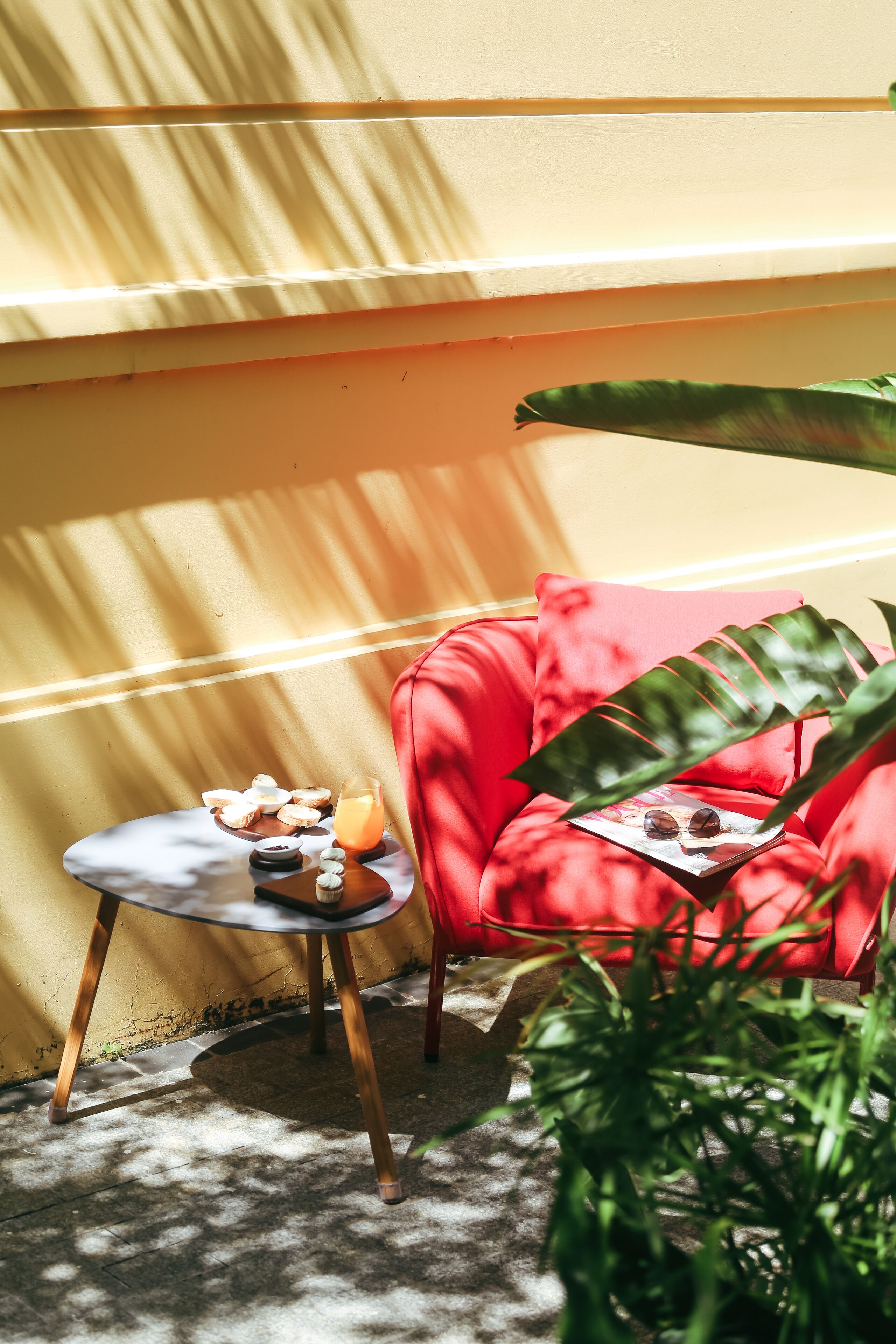 A red chair with a magazine and sunglasses, a small table with snacks and a drink, and a large green plant.