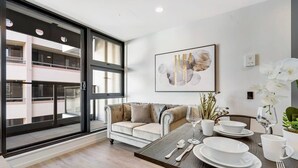 A modern living room with a sofa, dining table, and a large window overlooking a building. - CBD Modern Studio Apartment (Auckland)