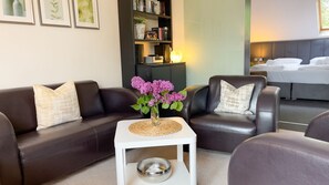 A modern living room with a brown leather sofa, a white side table with a decorative bowl, and a vase with purple flowers. A bookshelf with books and decorative items is in the background. - Haus Leuchtfeuer - Haus Leuchtfeuer - Bright vacation rental near the beach with (Cuxhaven)