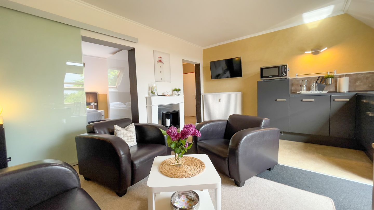 A compact living space with a sofa set, a small table with a vase of flowers, a kitchen area with appliances, and a television on the wall.