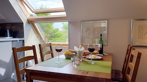 A dining area with a table set for two, featuring a bottle of wine, glasses, a bowl of fruit, and a plate of cheese. The room has a skylight, a framed picture on the wall, and a view of greenery outside.