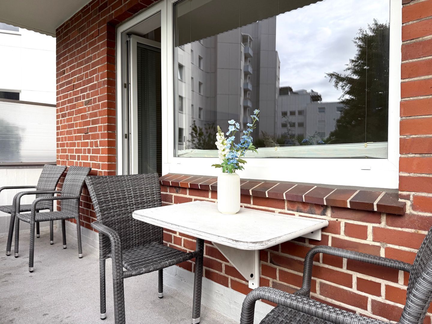 Ein Sitzbereich im Freien mit Korbstühlen und einem kleinen Tisch, ein Fenster mit Blick auf Gebäude und ein Blumentopf auf der Fensterbank.