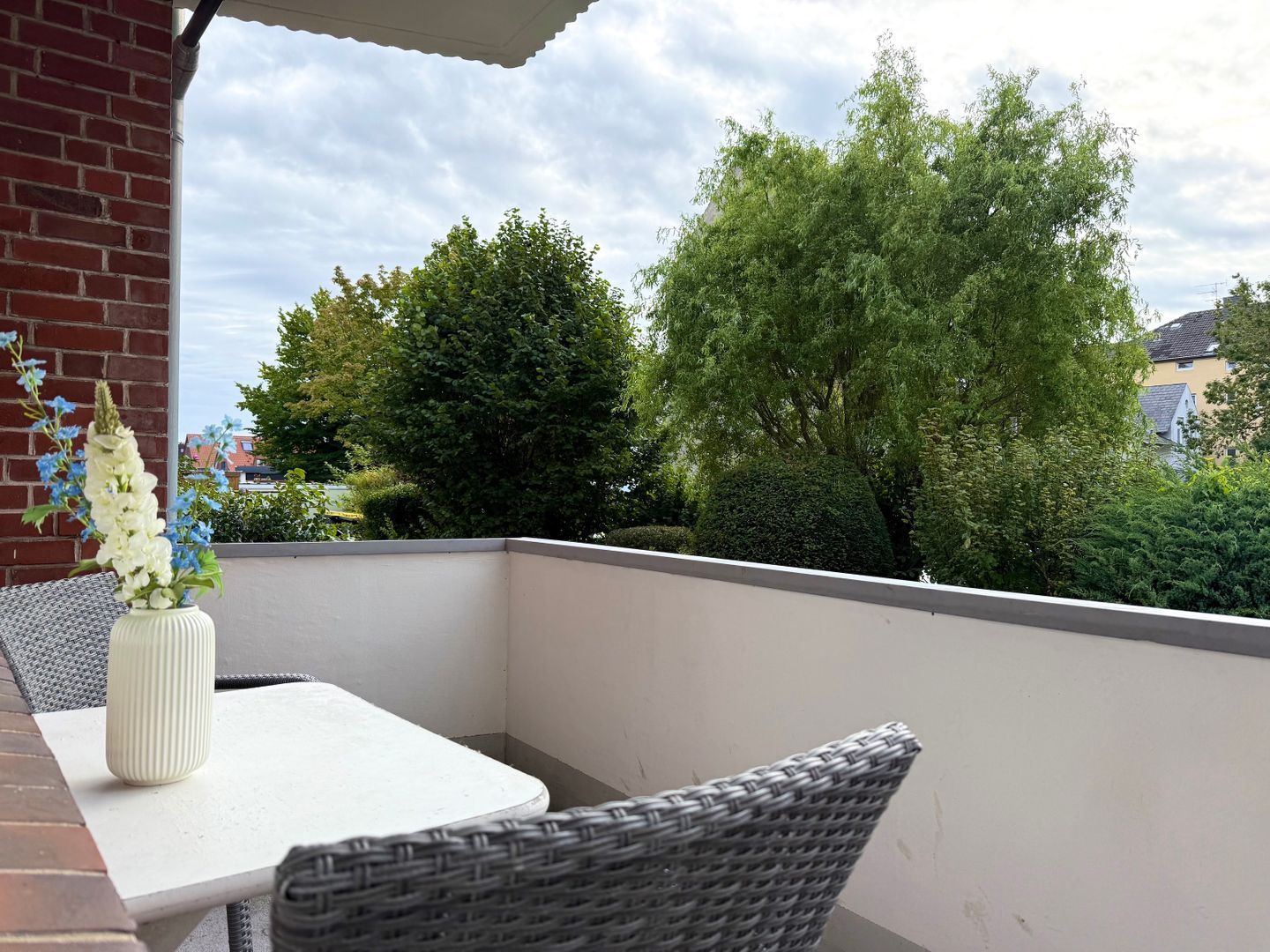 Ein Balkon mit einem weißen Tisch und Korbstühlen, mit Blick auf eine grüne Landschaft mit Bäumen und einem Gebäude im Hintergrund.