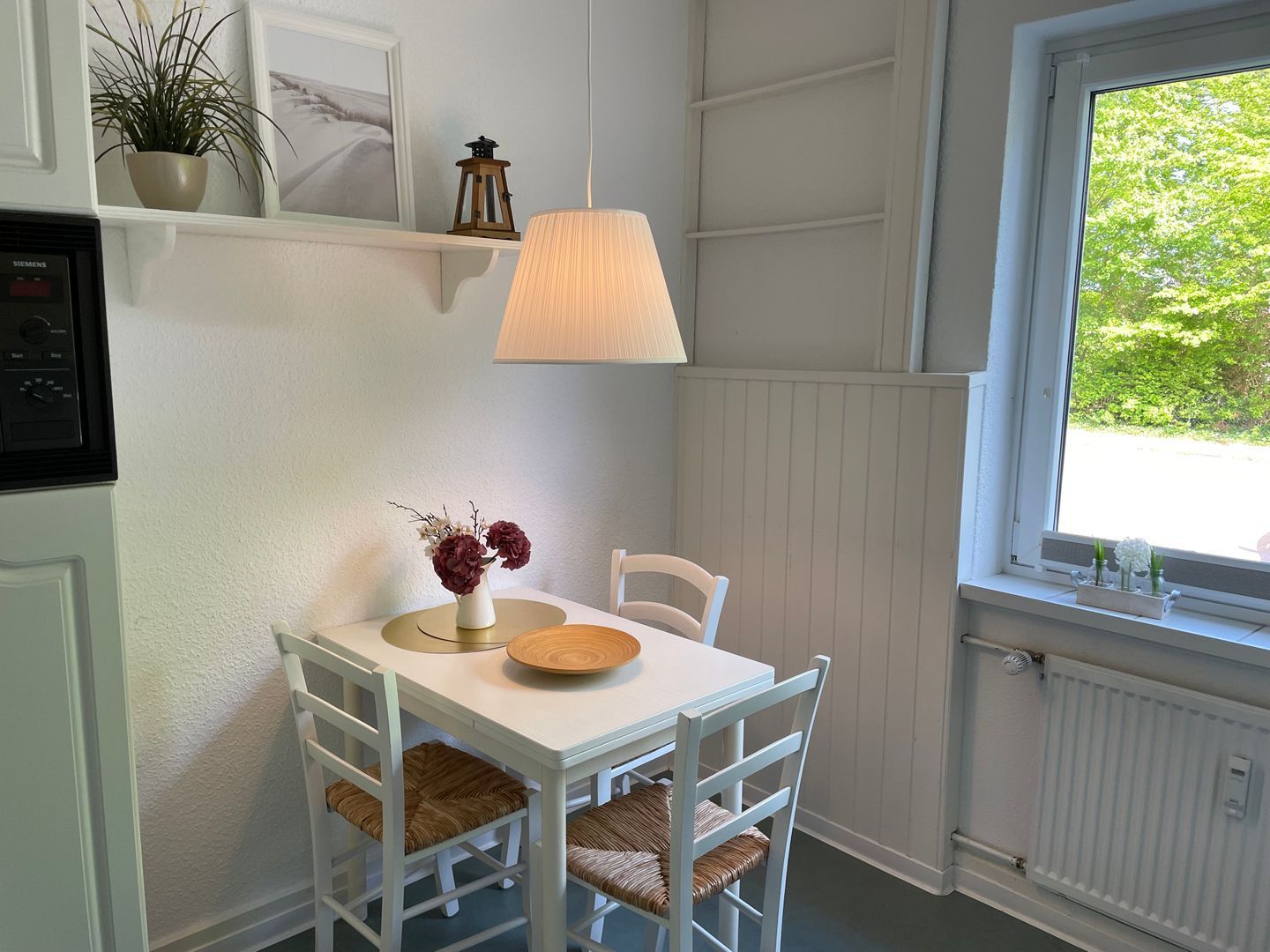 Ein kleiner Essbereich mit einem weißen Tisch und Stühlen, einer Lampe, einer Vase mit Blumen und einem Fenster mit Blick auf Grünflächen.