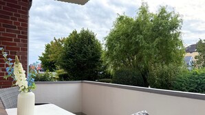 Un balcone con un tavolo bianco e sedie in vimini, con vista su un paesaggio verde con alberi e un edificio sullo sfondo.