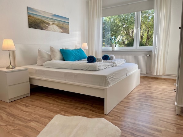 A modern bedroom with a large bed, white bedding, and a view of trees through a window.