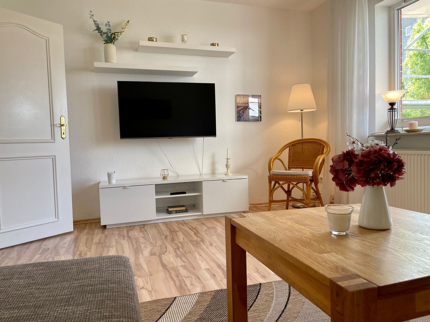 A modern living room with a flat-screen TV mounted on the wall, a wooden dining table, a wicker chair, and a vase with flowers.